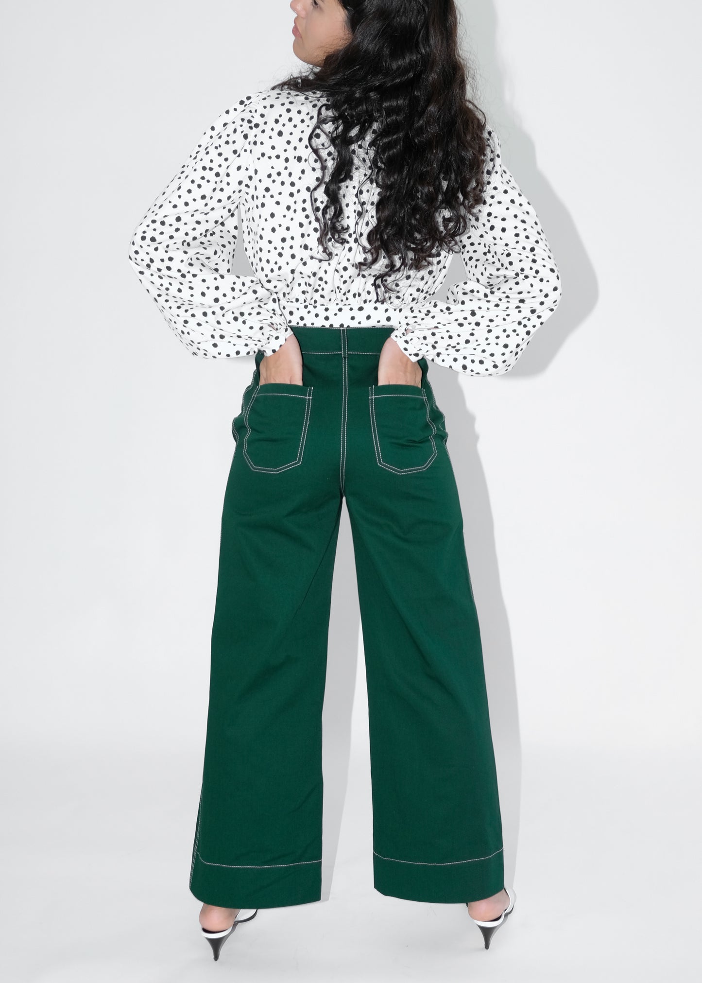 Back view of woman wearing pine green trousers and white blouse with black polka dots