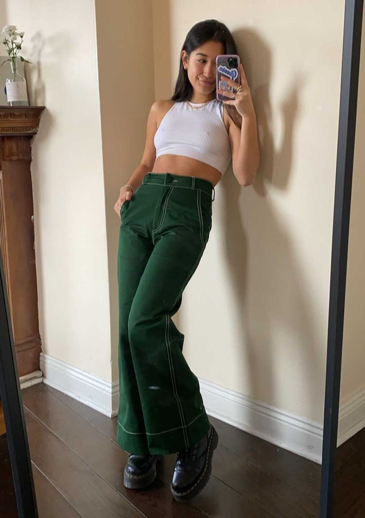 Woman wearing green trousers and white tank top taking picture from a mirror